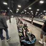 People wearing helmets sit in a line of go-karts on an indoor track, with others standing nearby. The area is well-lit and spacious.