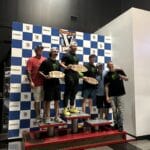 Six men pose on a winners' podium in front of a checkered backdrop, holding award plaques, with one man standing on the tallest platform.