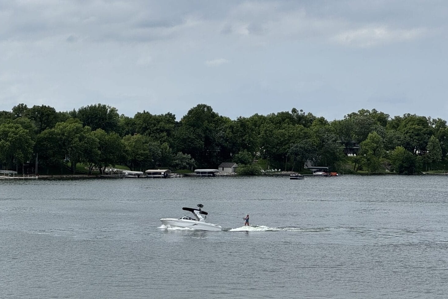 A person is wakeboarding behind a white boat on a lake, with another boat nearby and trees lining the shore under a cloudy sky.