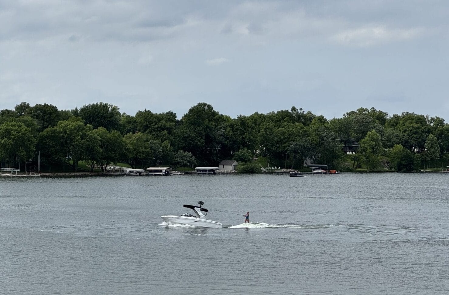 A person is wakeboarding behind a white boat on a lake, with another boat nearby and trees lining the shore under a cloudy sky.