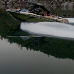 A green Centurion speedboat with several people onboard moves quickly across calm water, leaving a white wake behind. Rocky shoreline and trees are reflected in the lake.