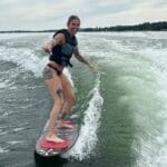Person wearing a life vest and swimsuit is wakesurfing on a lake, balancing on a surfboard behind a boat, with trees and cloudy sky in the background.