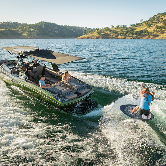 A motorboat pulls a person on a wakeboard across a lake, with three people sitting on the boat and hills in the background.