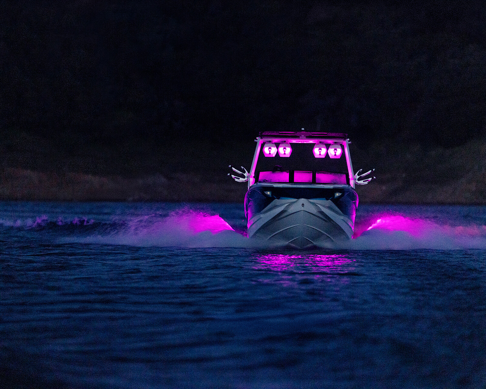 A Nv243 Wake Boat glides through dark water at night, its bright purple lights illuminating the exterior and reflecting on the water’s surface.