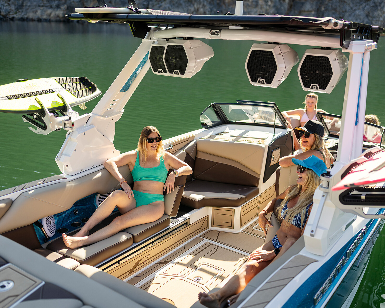 Four people relax and chat on a modern Centurion Nv243 Wake Boat in calm, green water, with wakeboards visible on board.