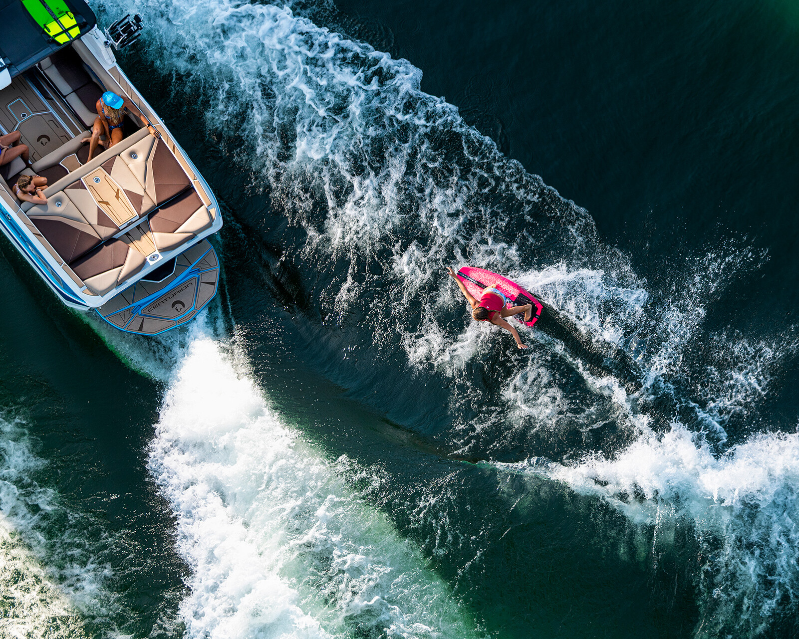A person rides a pink wakeboard behind a Centurion Nv243 on open water, creating white waves, while two people sit inside the boat.