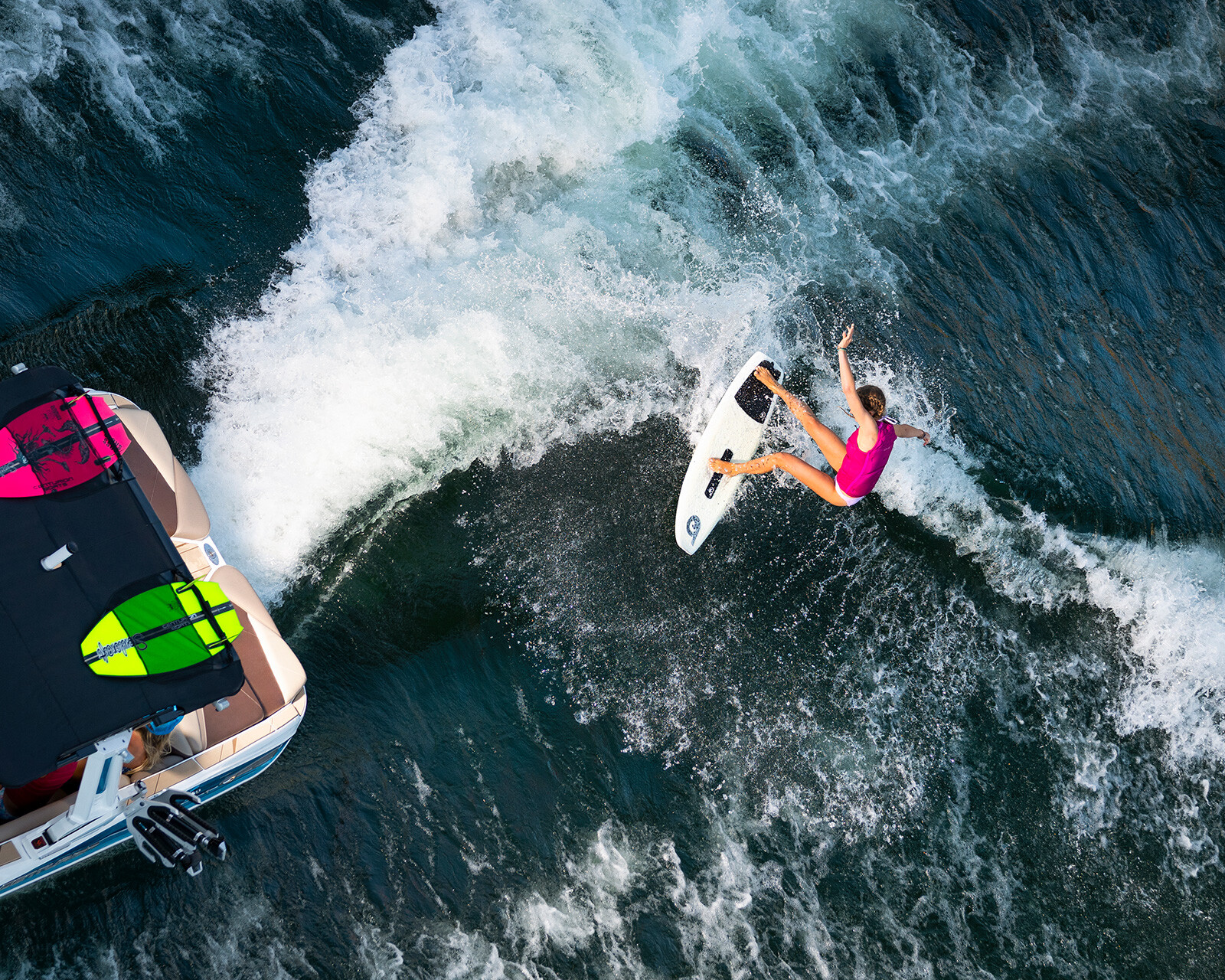 A person in a pink outfit is wakesurfing behind a Centurion Nv243, which has two surfboards on its roof and creates perfect waves in the water.