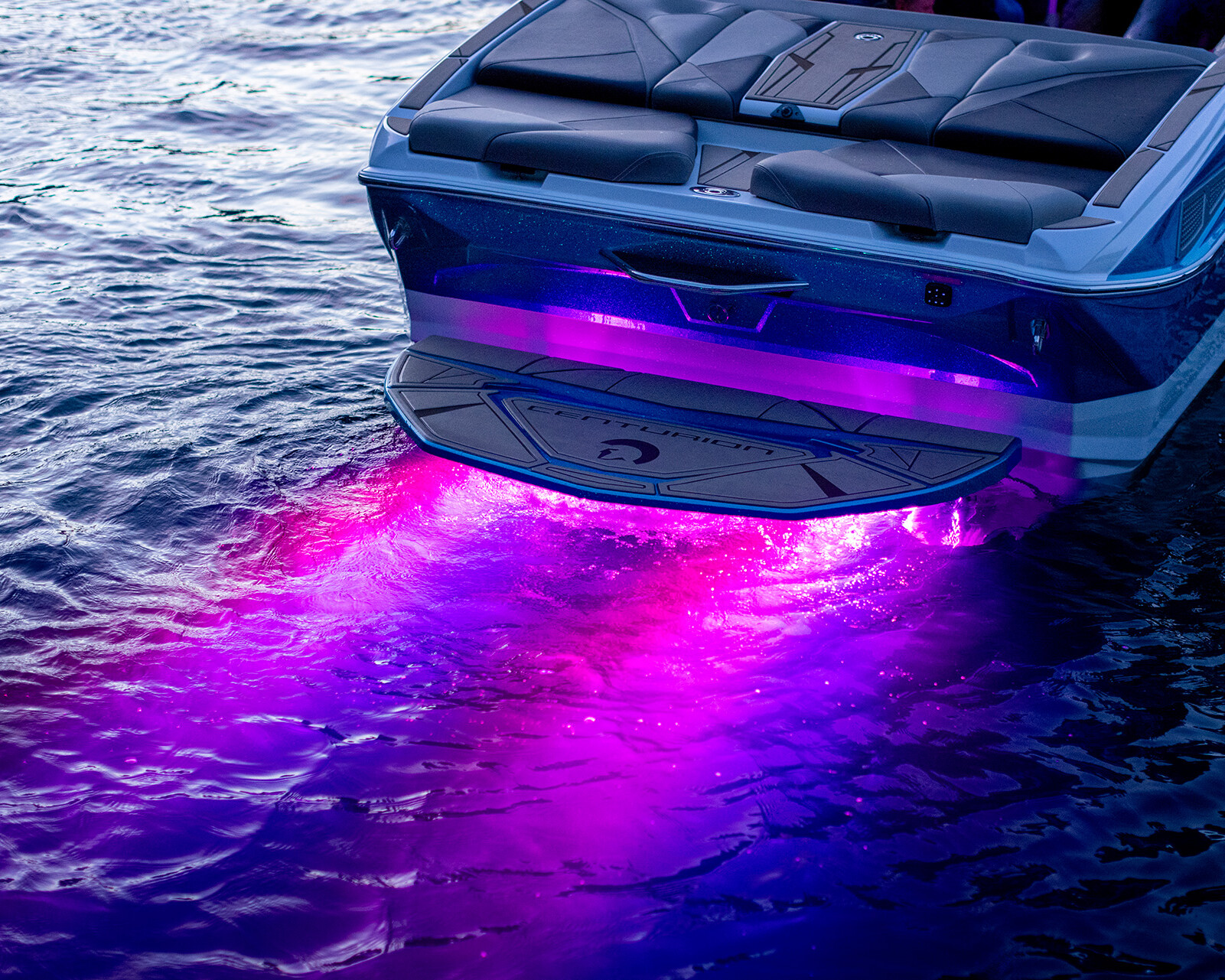 The back of a Centurion Nv243 Wake Boat with purple underwater LED lights illuminates the water at dusk.