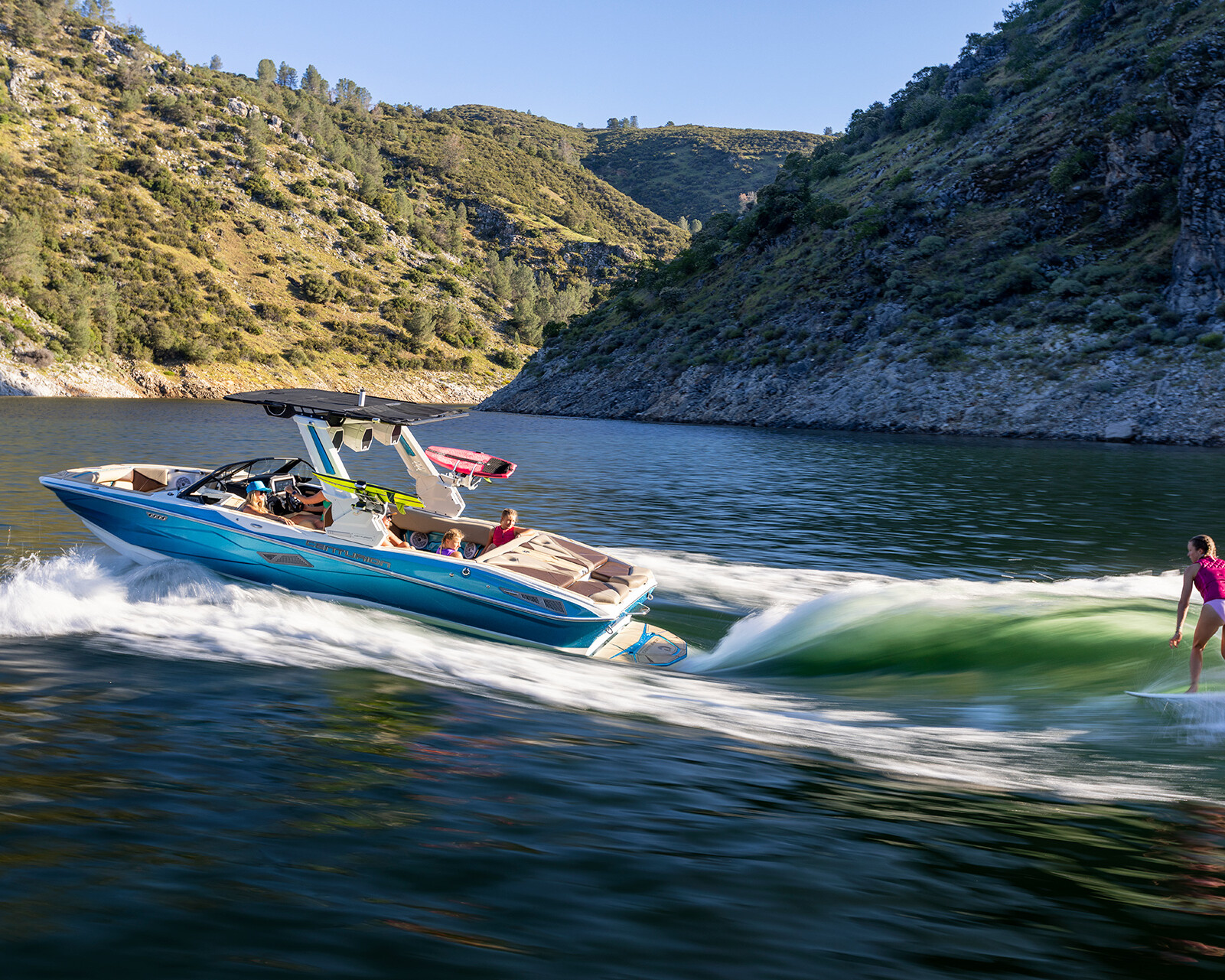 A person in a pink outfit surfs behind a Centurion Nv243 wake boat on a lake surrounded by green hills and rocky slopes.