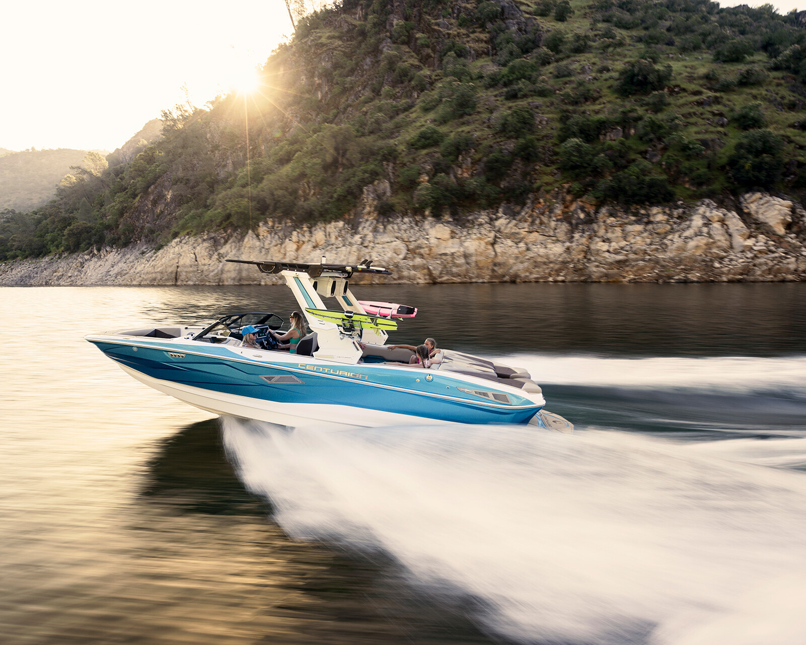 A blue and white Centurion Nv243 speedboat moves quickly across a lake near rocky, tree-covered hills at sunset, leaving a wake behind it.