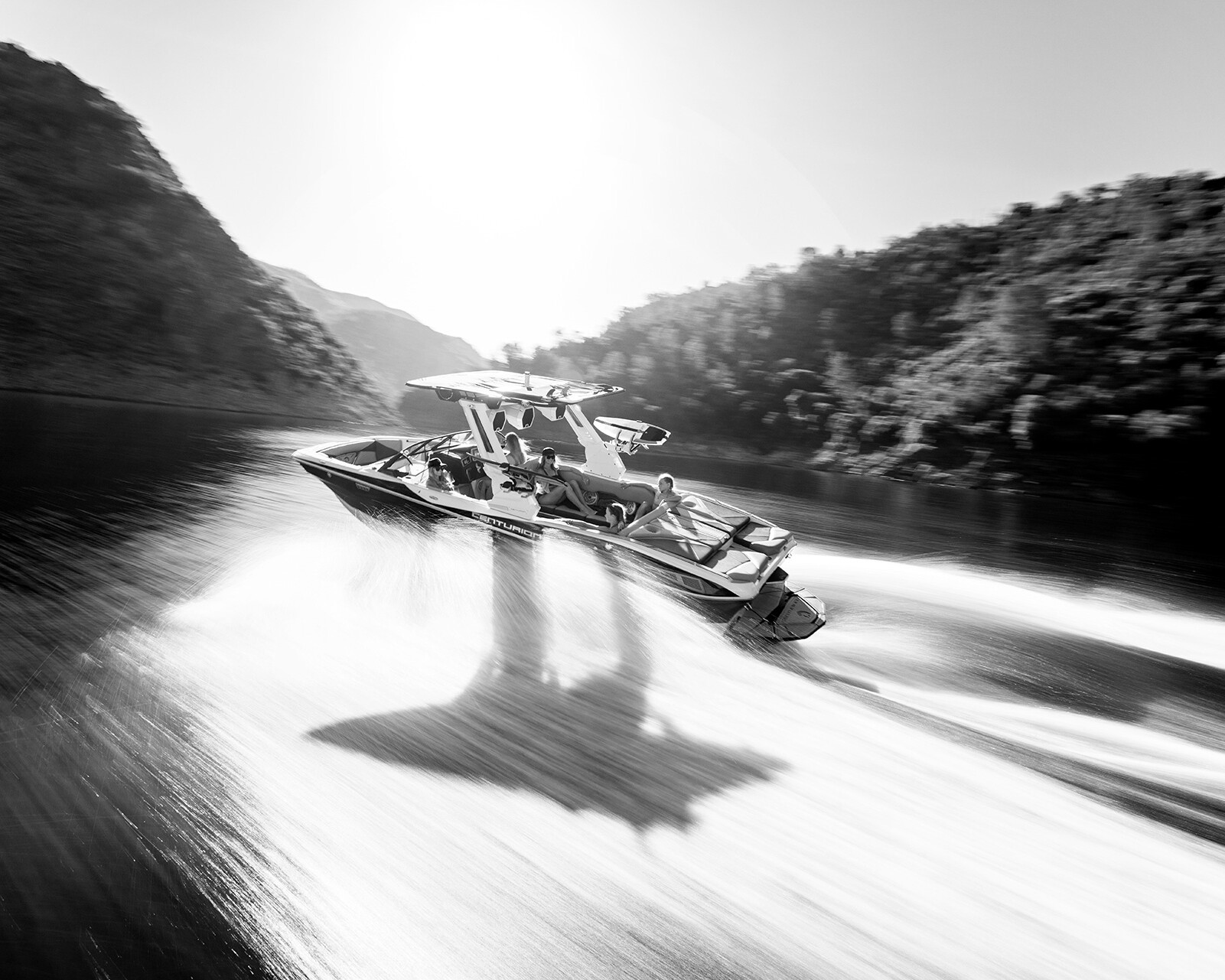 A Centurion Nv243 Surf Boat speeds across a river or lake, leaving a large wake behind, with hills rising on either side under bright sunlight.