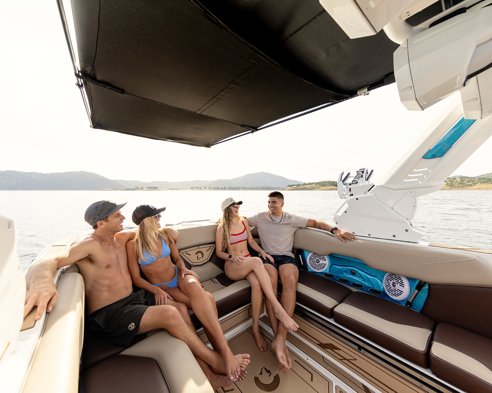 Four people in swimwear sit and talk on the seating area of a Centurion Nv243, with a lake and hills visible in the background.
