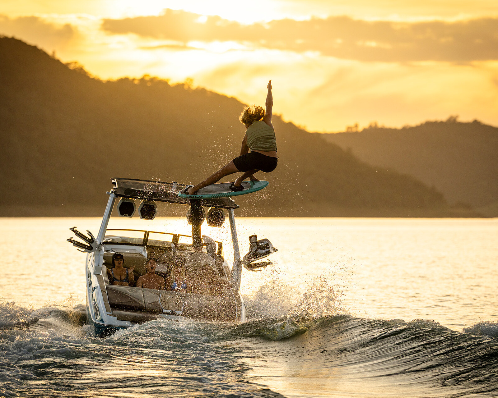 A person wake surfing behind a Centurion Nv243 with several passengers on a lake at sunset, surrounded by hills.