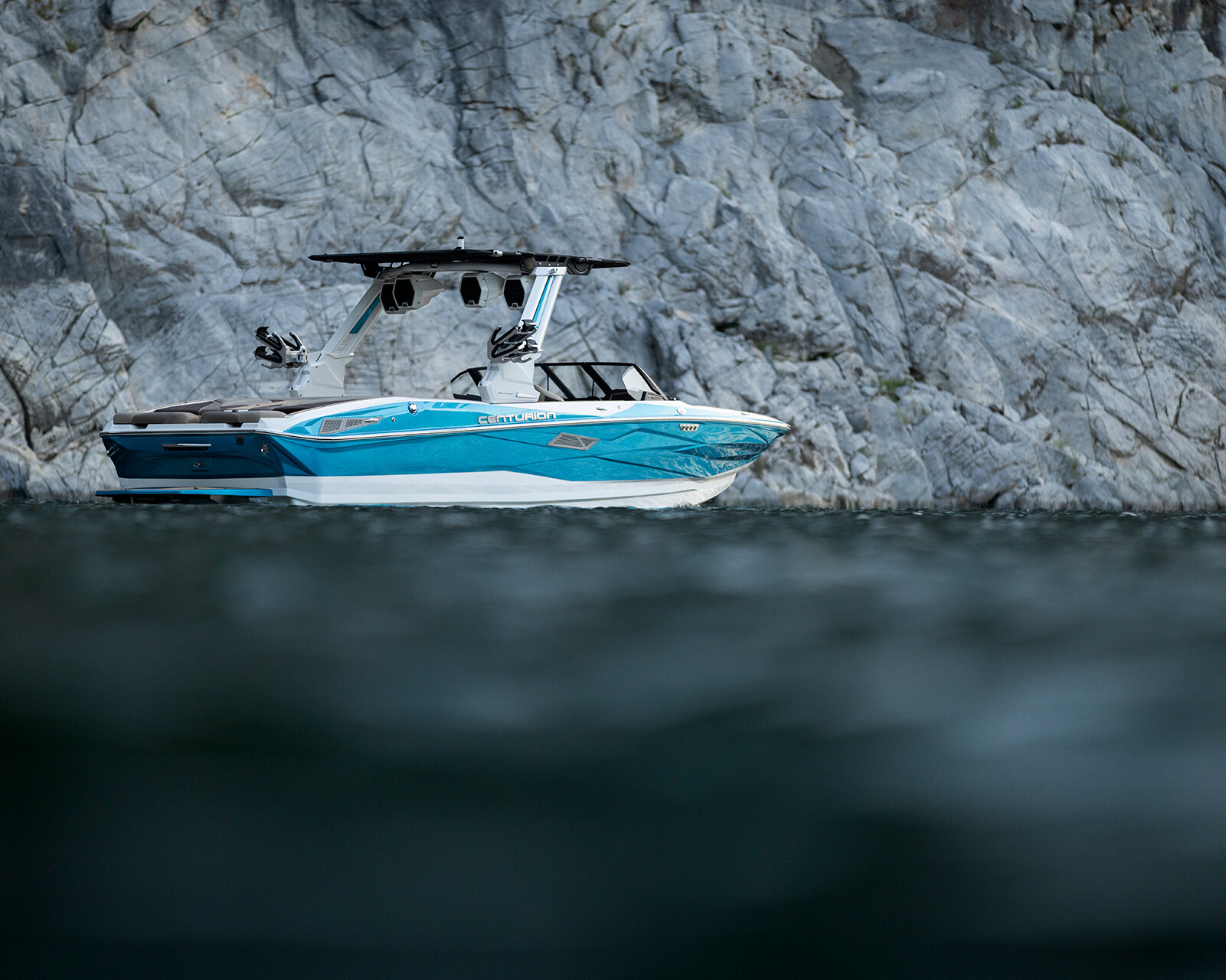 An Nv243 Wake Boat in blue and white is stationary on the water near a rocky, gray cliff.
