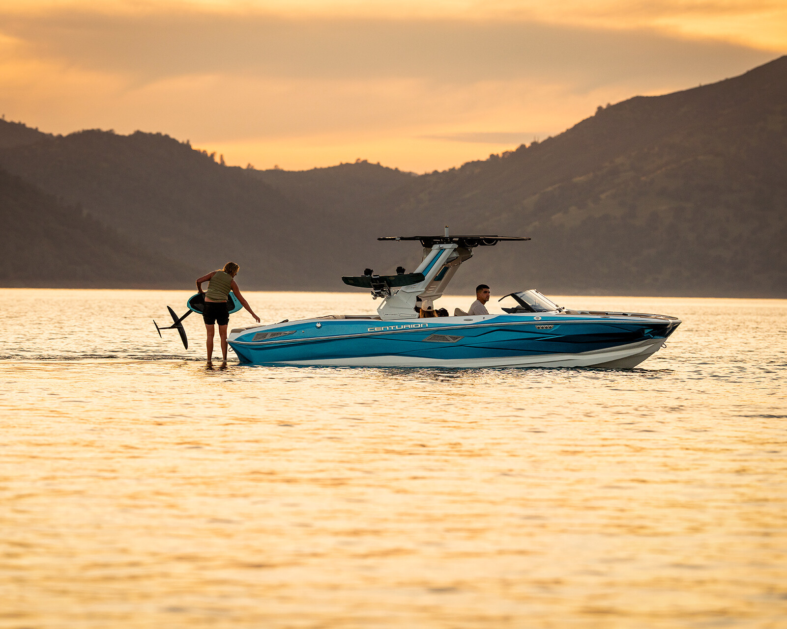 A person rides a hydrofoil surfboard behind a Centurion Nv243 on a calm lake at sunset, with hills visible in the background.