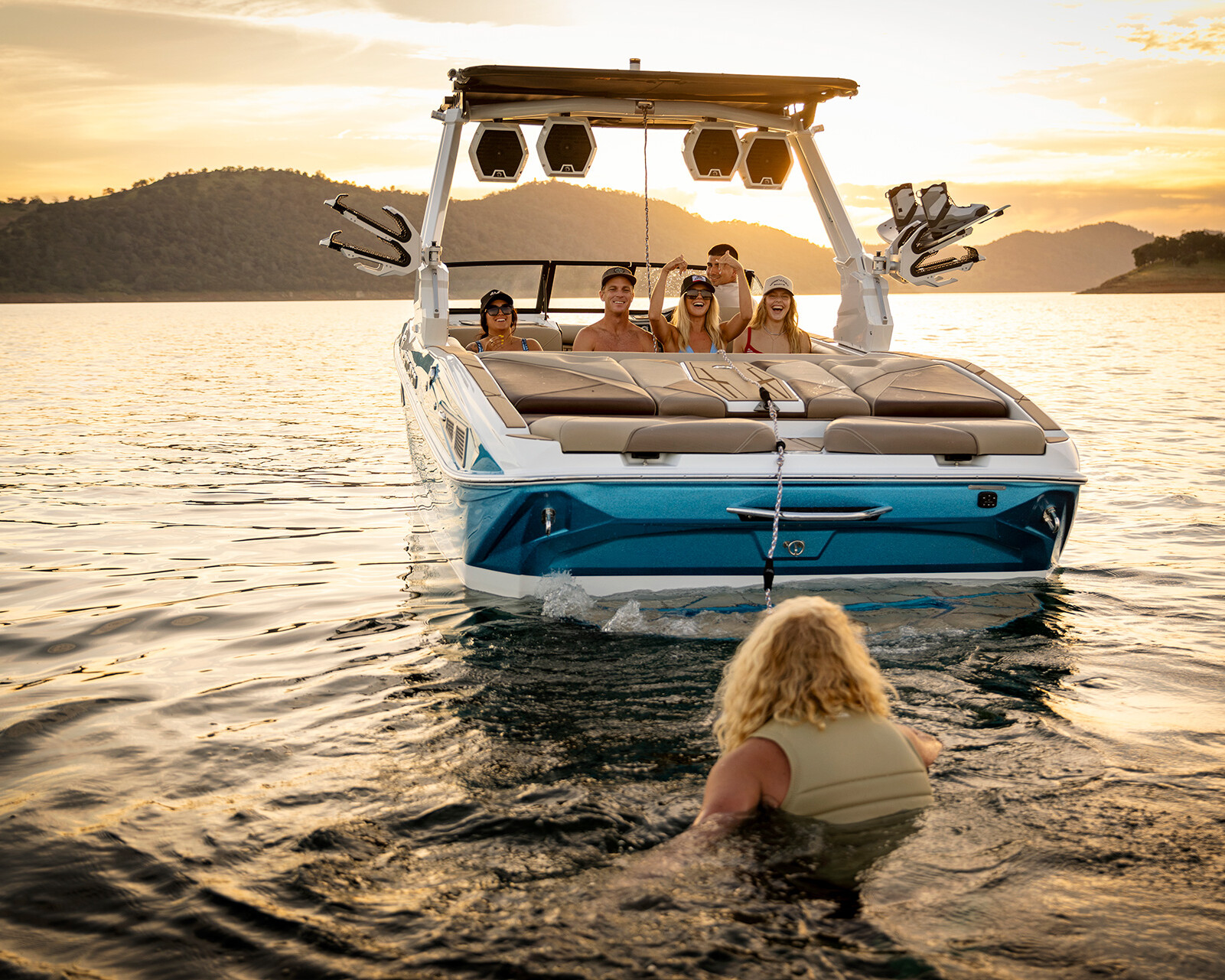 A person in a life vest floats in the water behind a blue and white Centurion Nv243 wake boat, with five people seated onboard at sunset.