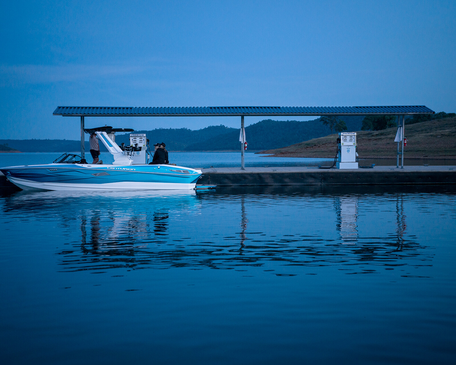 A white and blue Centurion Nv243 Wake Boat is docked at a lakeside fueling station, with two gas pumps under a metal canopy in the early evening.