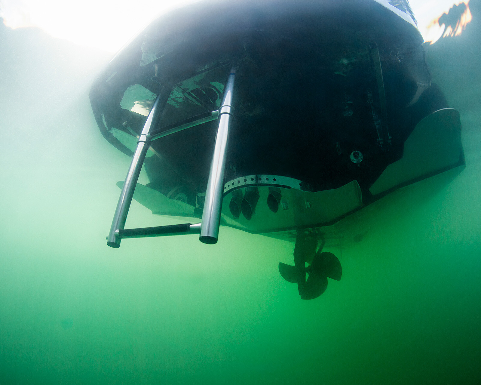 Underwater view of the stern of an Nv243 Surf Boat, showing a metal swim ladder and a propeller submerged in greenish water.