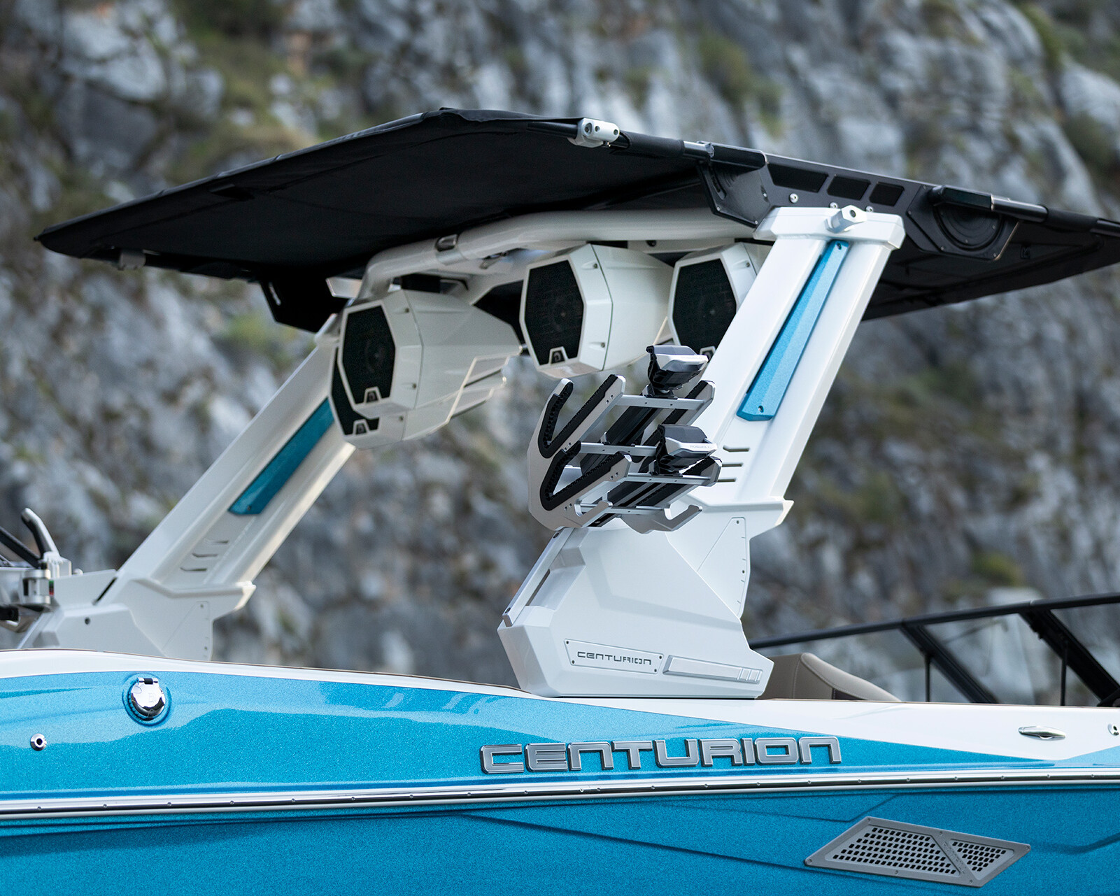 A close-up of a blue Centurion Nv243 surf boat highlights its wakeboard tower with speakers, racks, and a black sunshade, set against rocky cliffs in the background.