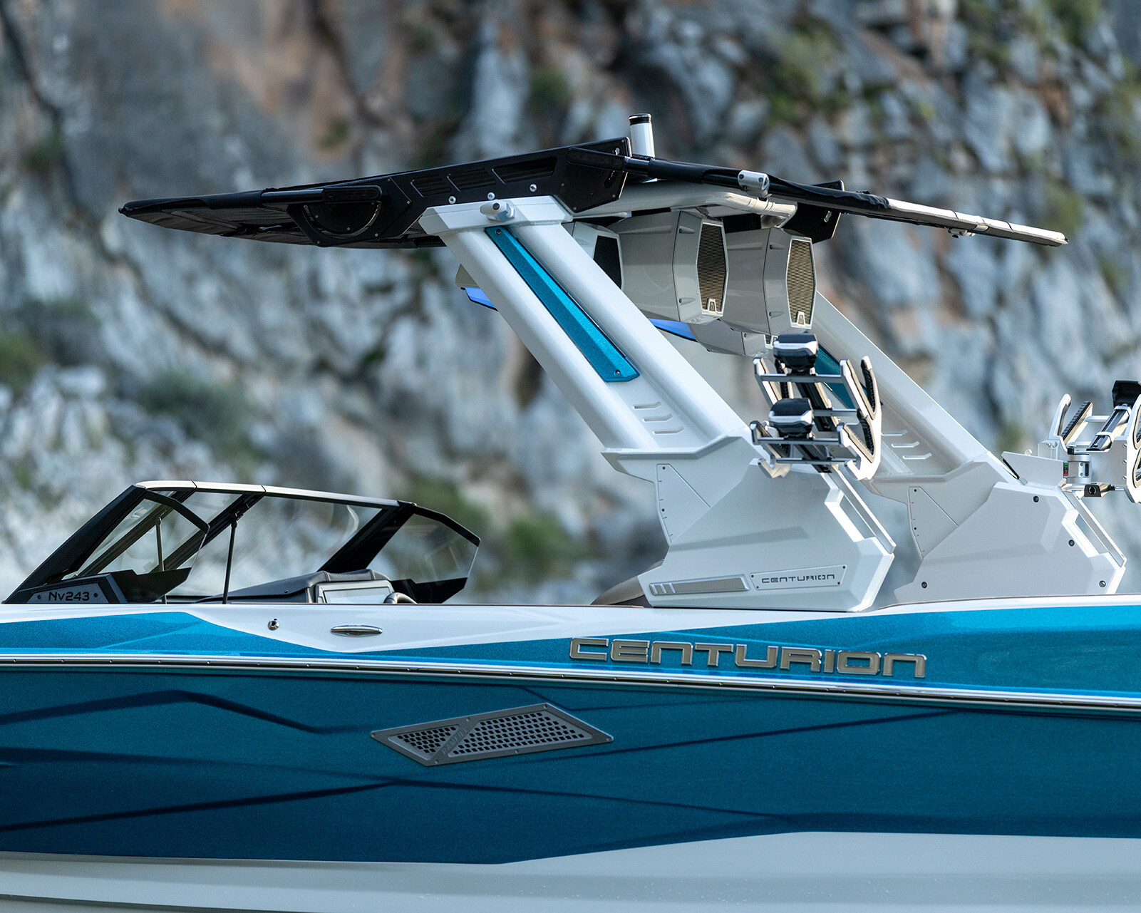 A blue and white Centurion Nv243 Wake Boat with a black wakeboard tower and two wakeboard racks, shown near a rocky shoreline.