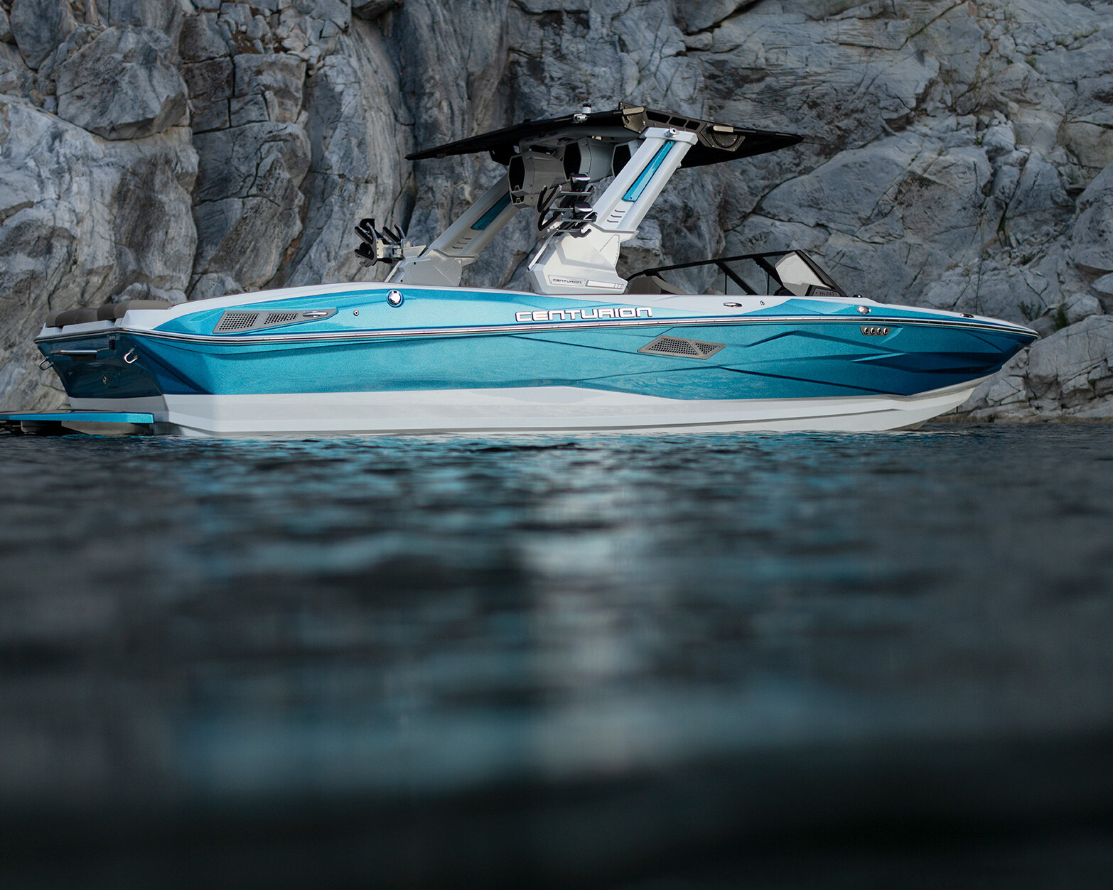 A blue and white Centurion Nv243 Wake Boat floats on calm water near a rocky cliff shoreline.