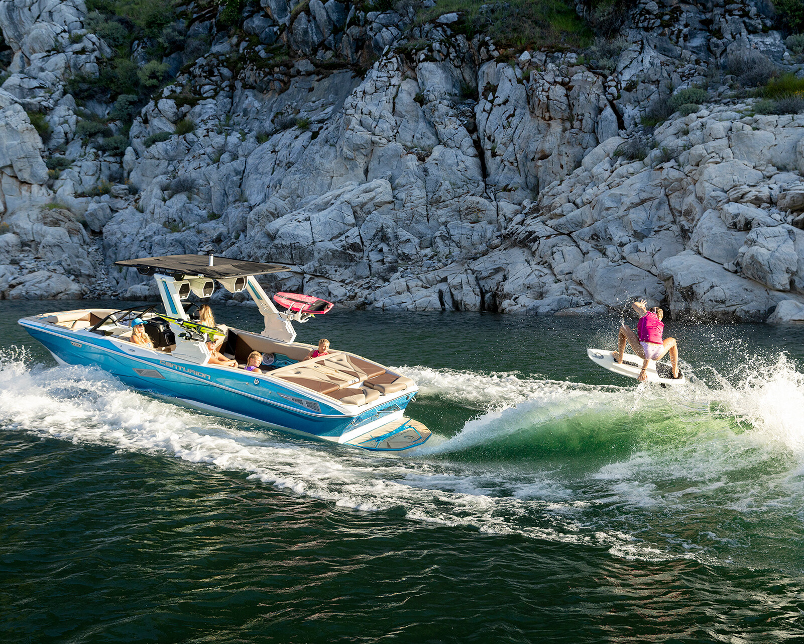A Nv243 Wake Boat pulls a person on a wakeboard across green water near a rocky shoreline, with several passengers onboard watching.