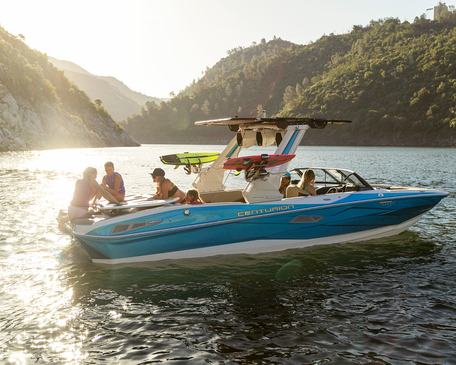 A group of people sit and relax on a blue Centurion Nv243 Surf Boat on a lake, surrounded by hills and sunlight reflecting on the water.