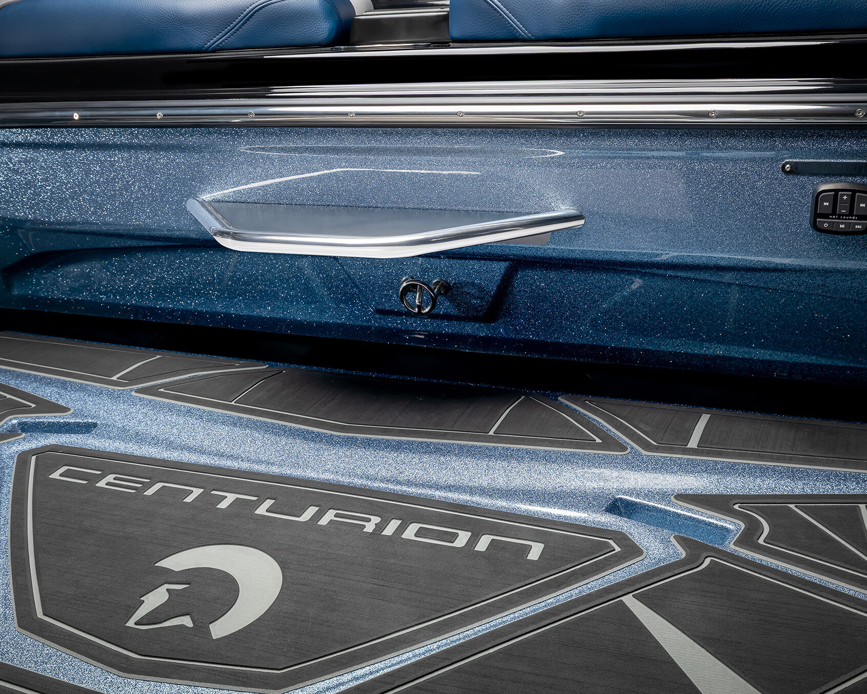 Close-up view of a Centurion Nv243 Wake Boat’s interior, featuring a blue metallic side panel, silver handle, control switch, and branded non-slip flooring with the Centurion logo.