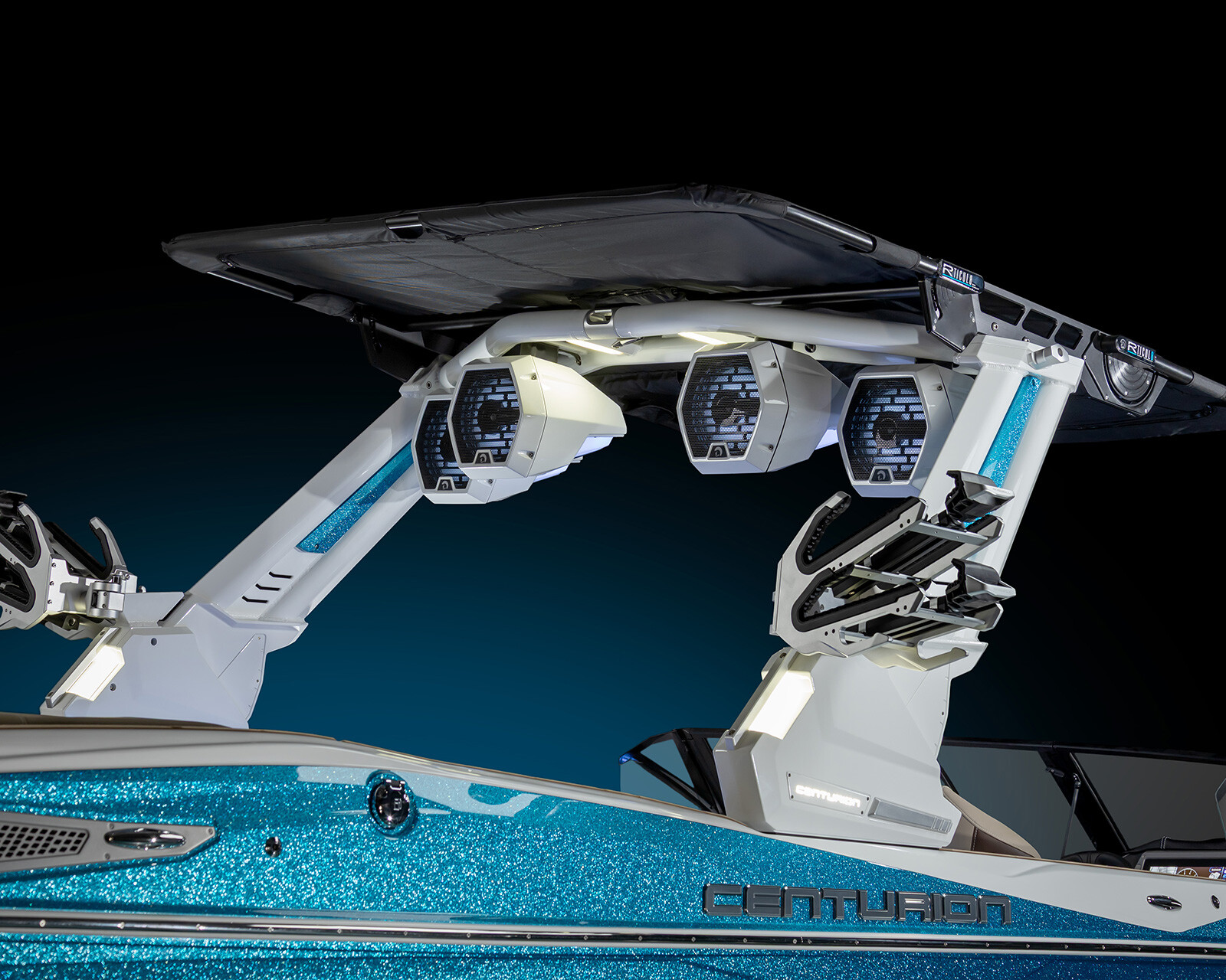 A close-up of an Nv243 Surf Boat's wakeboard tower with mounted speakers, wakeboard racks, and a sunshade, showcasing a metallic blue and white finish.