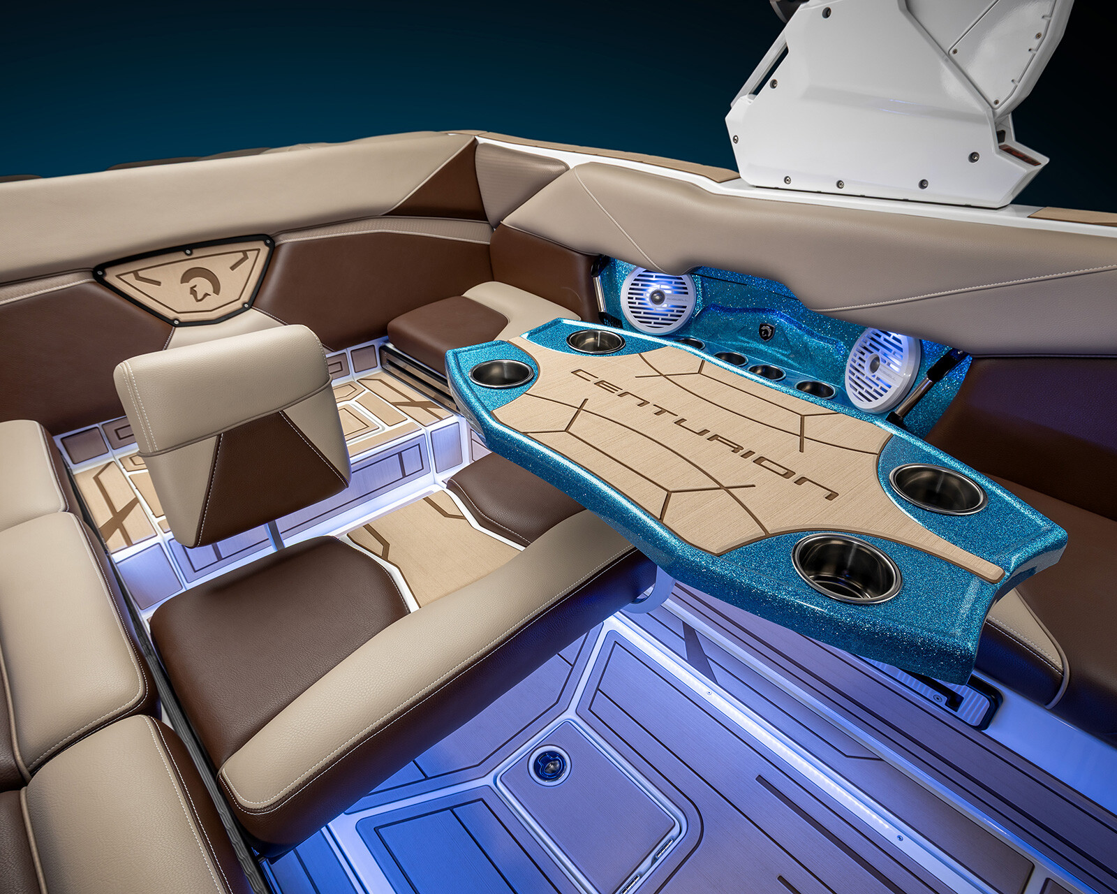 Luxurious Centurion Nv243 surf boat interior with brown and tan cushioned seating, a blue Centurion-branded table with cup holders, built-in speakers, and blue accent lighting for an elevated on-water experience.