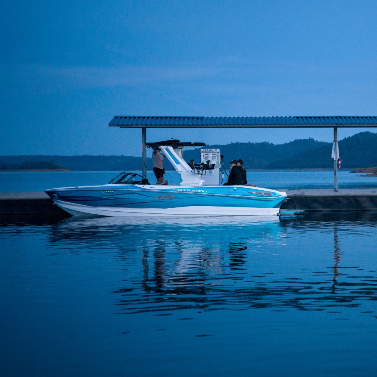 A blue and white speedboat is docked beside a covered fuel station on a calm lake at dusk, with hills visible in the background.