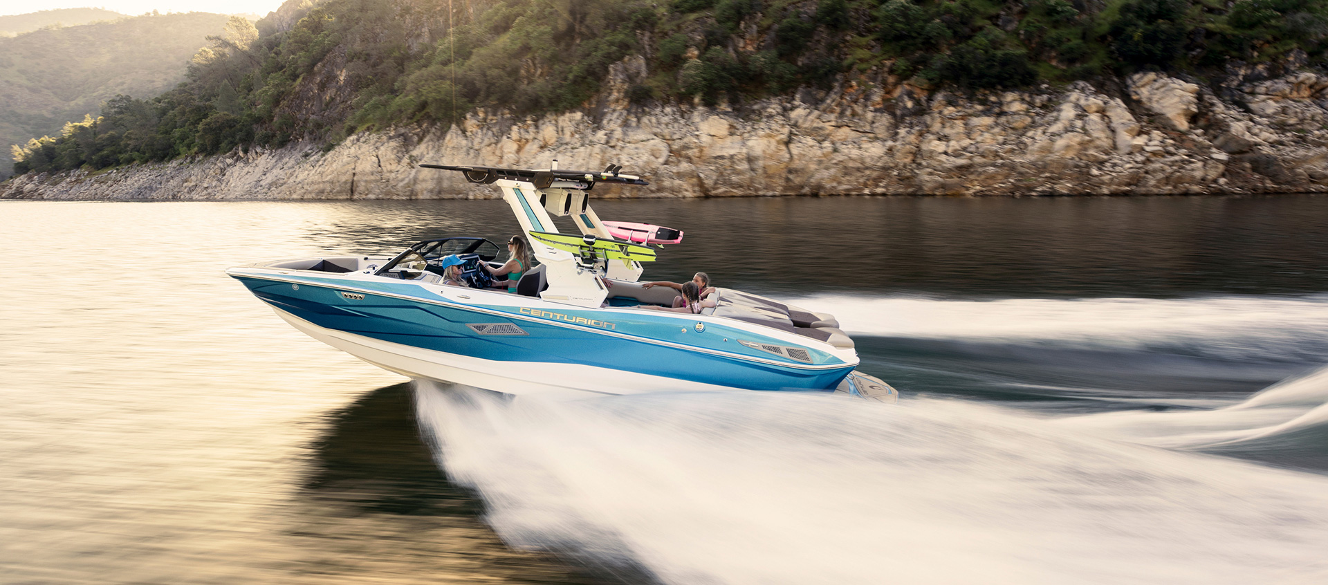 A blue and white Nv243 Wake Boat with people onboard speeds across a lake, creating waves, with a rocky, tree-lined shore in the background.