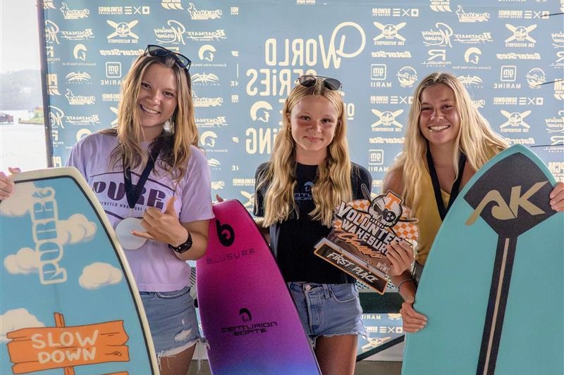 Three young women stand holding skimboards and a trophy in front of a branded backdrop at the 2025 Centurion WSWS Volunteer Wake Surf Classic Finals.