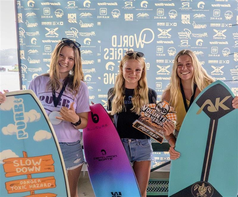 Three young women stand holding skimboards and a trophy in front of a branded backdrop at the 2025 Centurion WSWS Volunteer Wake Surf Classic Finals.