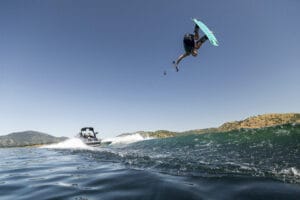 A person on a Fe22 wakeboard is airborne above the water while being towed by a boat on a sunny day, with hills visible in the background.