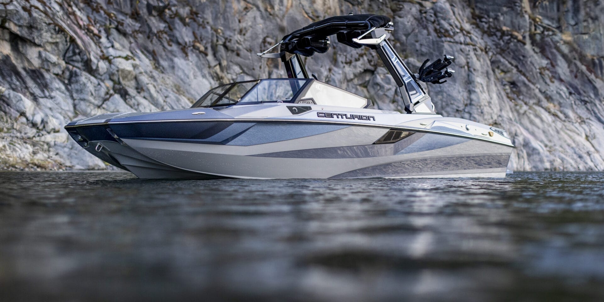 A modern 2026 Fe23 Centurion speedboat, a fearlessly evolved towboat, floats on calm water near a rocky cliff.