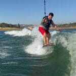 A person in a life vest and red shorts is wakesurfing on a lake behind a Centurion Boats vessel, balancing on a board while holding a rope, with waves and wooded hills in the background.