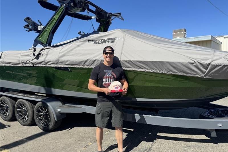 A person stands in front of a large covered Centurion Boat on a trailer, holding a small circular award. The scene is outdoors on a sunny day at a Ranger Road non-profit event.