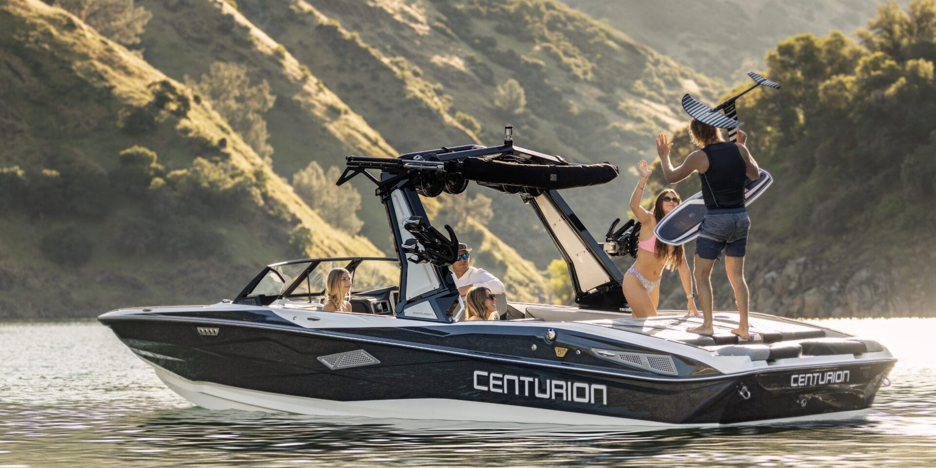 A group of people are on a 2026 Centurion motorboat, enjoying victory on the water near forested hills; one person proudly holds a wakeboard.