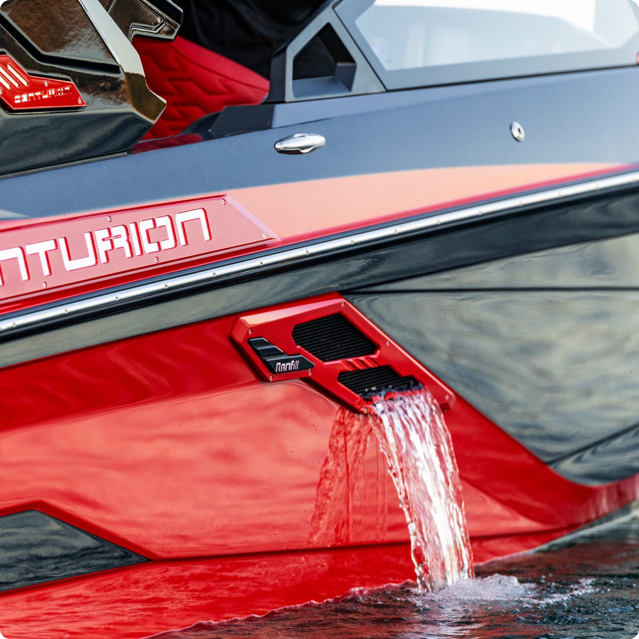 Close-up of a red and black Centurion boat with water flowing out of the Centurion RAMFILL vent on the side, partially reflected on the water's surface.