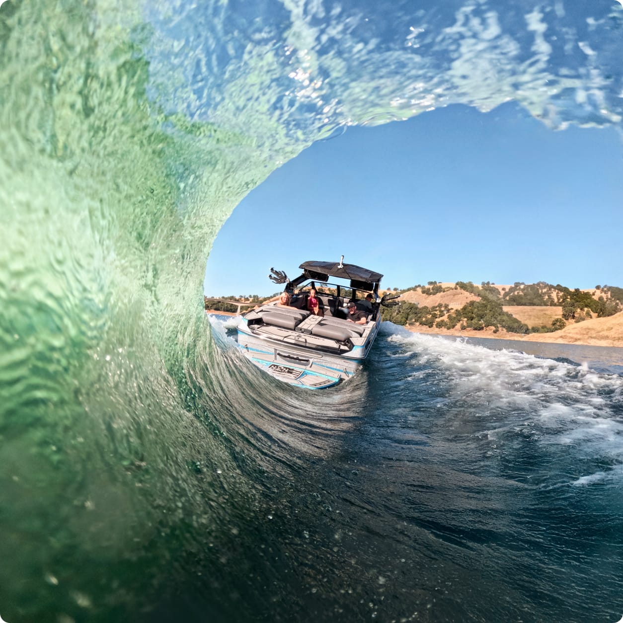A Centurion surf wave arches high on a sunny day, framing a boat with several people aboard and hills visible in the background.