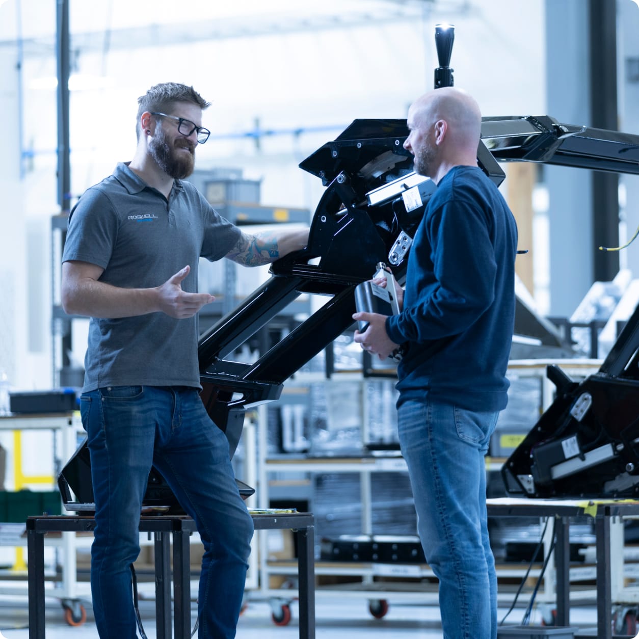 Two men stand and talk near industrial machinery in the bright Centurion Towers factory. One gestures while the other listens, both wearing casual clothes.