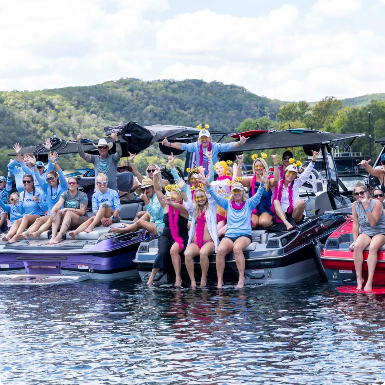 A group of people in colorful outfits and accessories sit on boats with the innovative Centurion Opti-V Hull, docked together on a lake, surrounded by hills and trees under a partly cloudy sky.