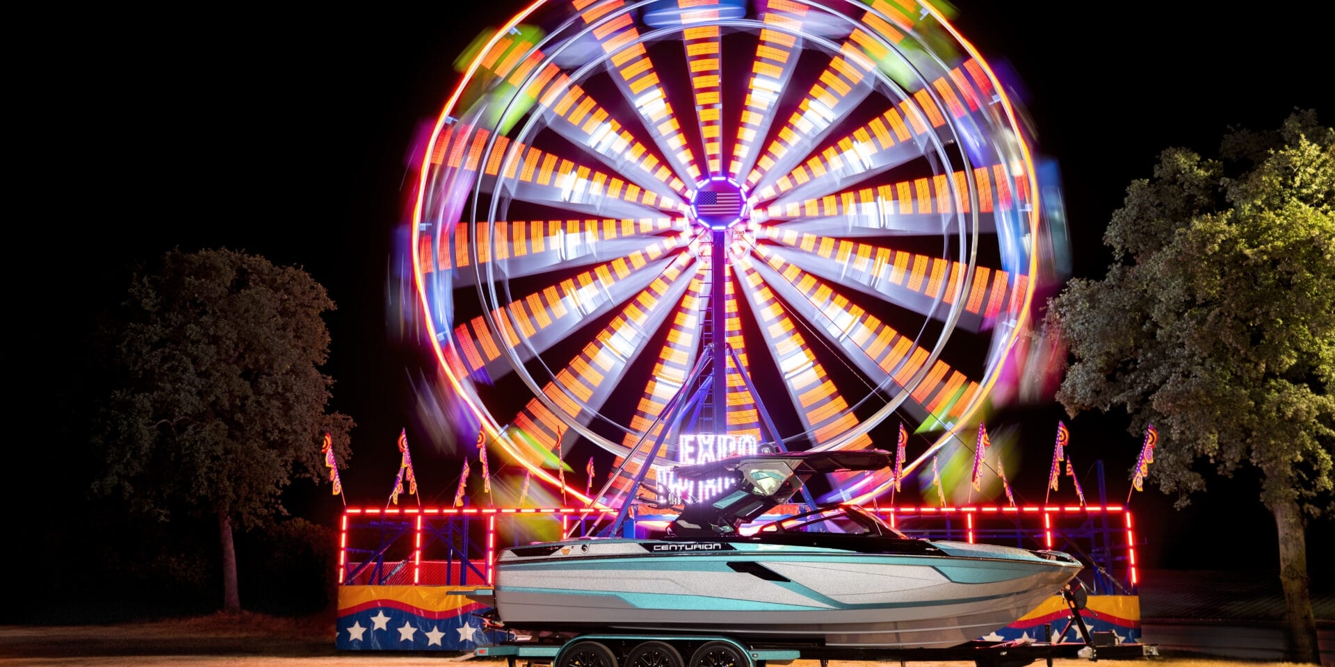 A brightly lit Ferris wheel spins at night behind a parked 2026 boat Ri245 on a trailer, with trees on either side and dry grass in the foreground—hinting at water sports adventures to come.