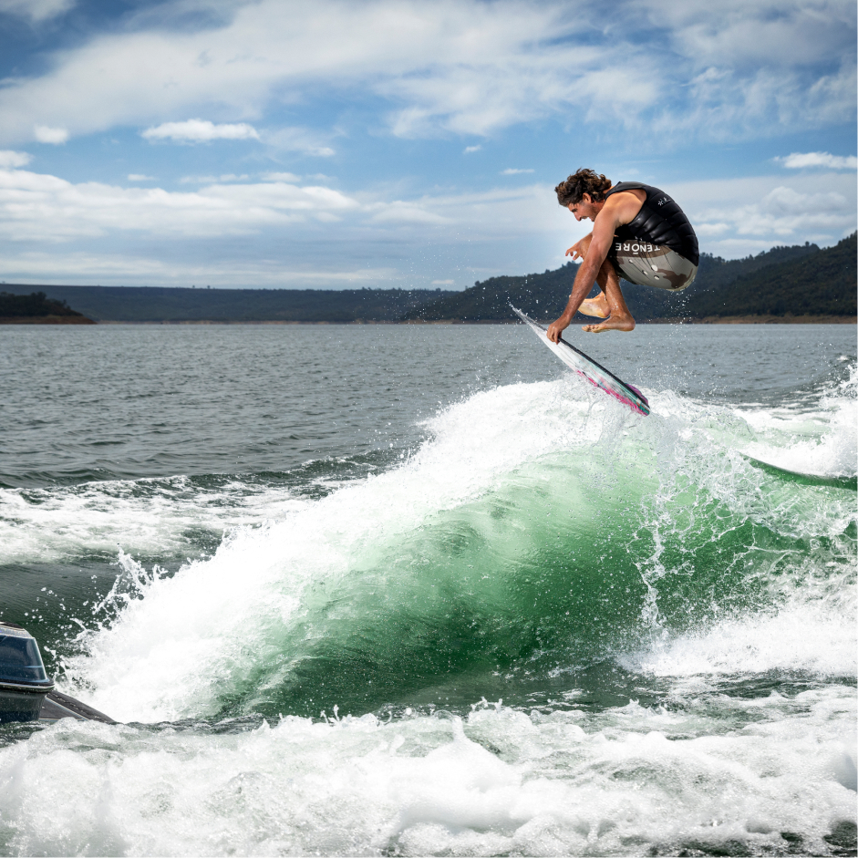 A man in a black vest is wake surfing on a green wave created by a boat, jumping in the air with a lake and hills in the background under a partly cloudy sky.
