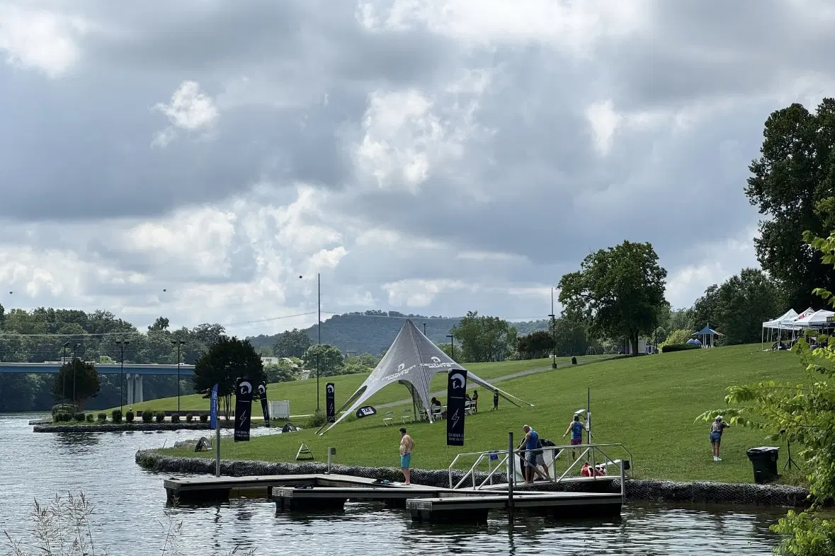 People stand on a dock and grassy shoreline near a lake, with the Volunteer Wake Surf Classic - Day 1 Recap banners and a white canopy tent set up on the lawn under a cloudy sky.