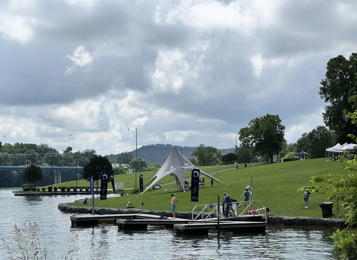 People stand on a dock and grassy shoreline near a lake, with the Volunteer Wake Surf Classic - Day 1 Recap banners and a white canopy tent set up on the lawn under a cloudy sky.