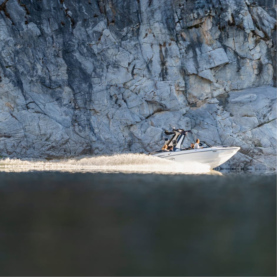 A white speedboat with an Opti-V Hull moves quickly across calm water near a large rocky cliff, leaving a visible wake behind it.