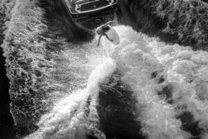 A person surfs on the wake behind a motorboat, with water splashing around them, captured from an overhead angle in black and white.