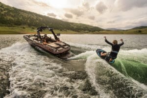 A person wakesurfs behind a motorboat on a lake surrounded by hills under a cloudy sky.